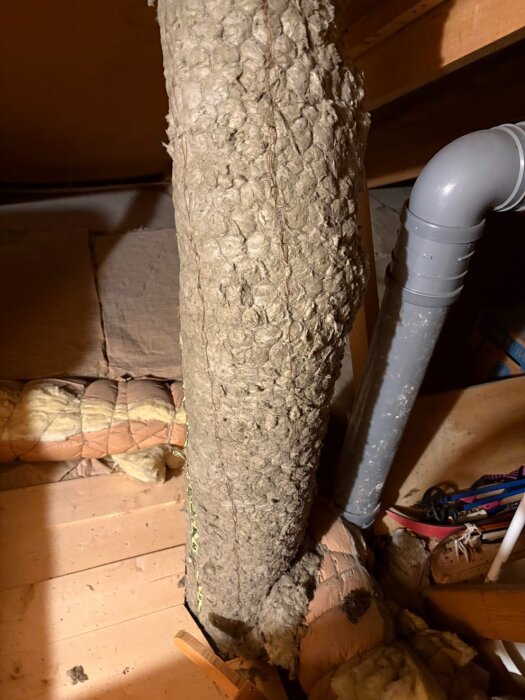 Isolerad ventilationskanal på vind med synlig mineralull och gammalt material, bredvid rör och andra byggkomponenter.