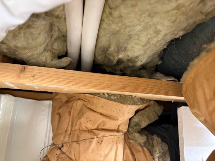 Isolering med mineralull runt rör och träreglar på vinden, med plast och pappersmaterial synligt. Diskuteras isoleringens skick för värmepump.