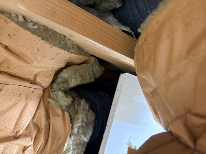 Isolering av avluftskanal på vind, med synlig mineralull och vad som kan vara en isoleringsstrumpa. Träbalk och byggplast syns också.