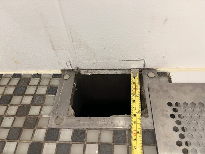 Golv med små fyrkantiga kakelplattor, en mätsticka mäter ett hål intill en vägg, bredvid ett metallgaller med hexagonmönster.