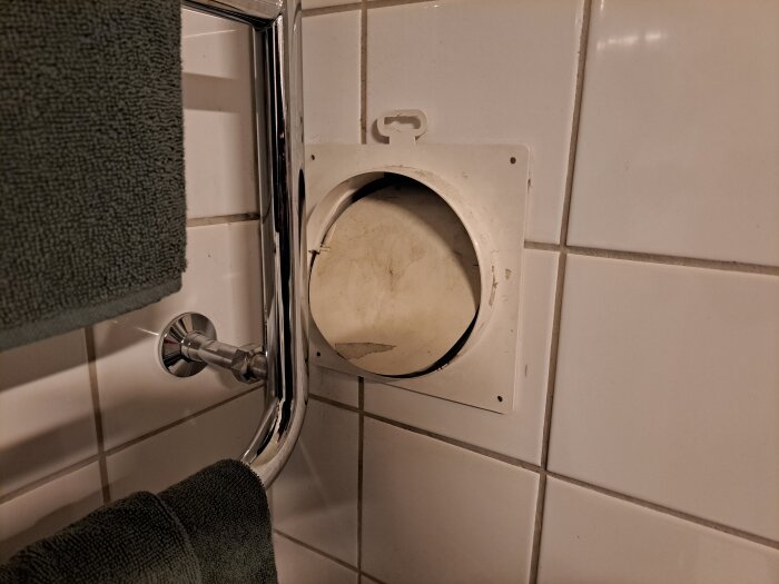 Öppen ventilationsöppning i ett kaklat badrum, med en handdukshängare och grön handduk till vänster.