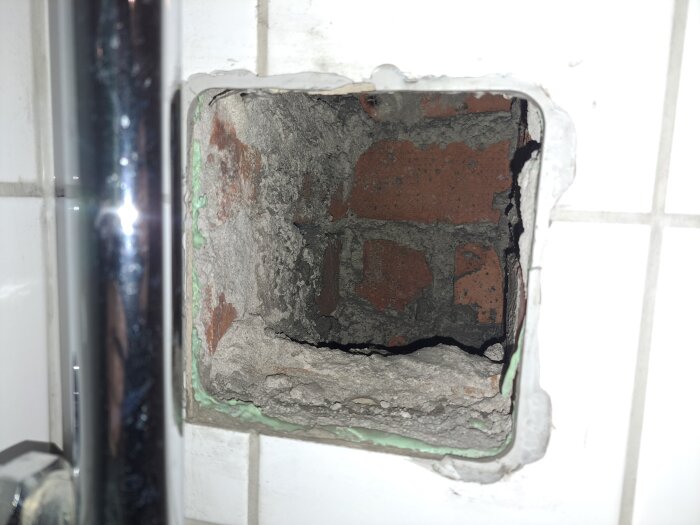 Hål i kaklat badrumsvägg med synliga tegelstenar och cement, del av kromad vattenrörsdetalj syns till vänster.