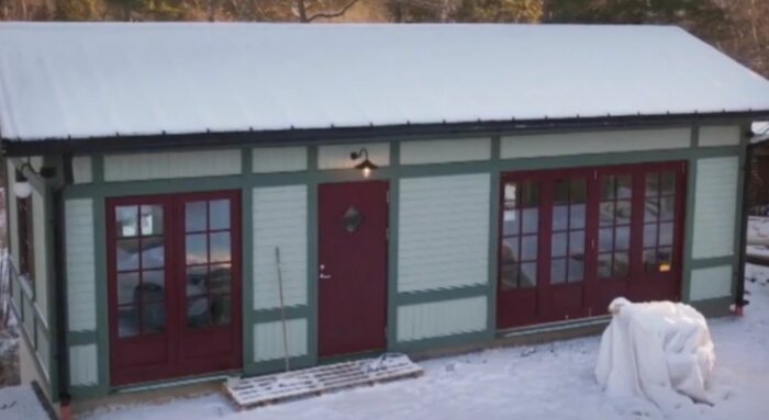 En komplementbyggnad i sekelskiftesstil med snötäckt tak. Röd dörr och fönster med gröna foder. Vintermiljö med snö på marken.