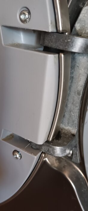 Närbild av trasiga skruvfästen och gångjärn på en vit maskinlucka, där skruvarna har lossnat, och luckan är delvis isärmonterad.