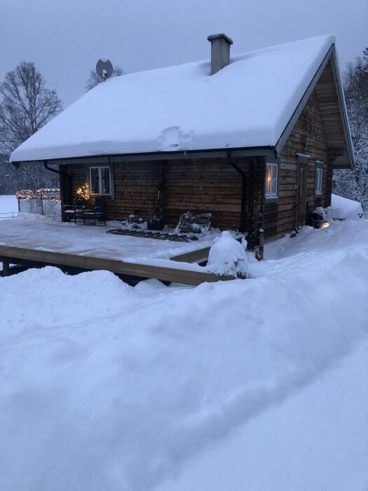 Trähus med snötäckt tak, snödrivor runt om, skottad altan, och snötyngda träd i bakgrunden under skymning.