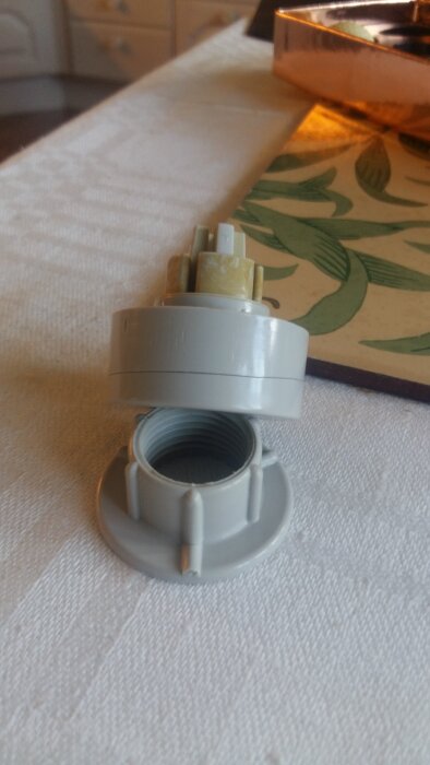 Grå plastadapter för rörkopplingar ligger på en bordduk bredvid en grönbladig underläggsbricka.