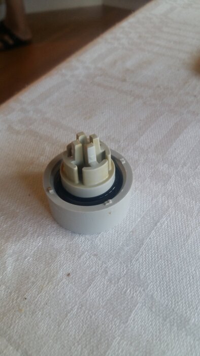Grå plastkomponent med flera utstickande delar, placerad på en vit tygyta.