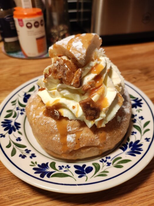 Gräddig semla med kolasås och kanderade valnötter på ett blommönstrat fat, bakgrund av köksdetaljer.