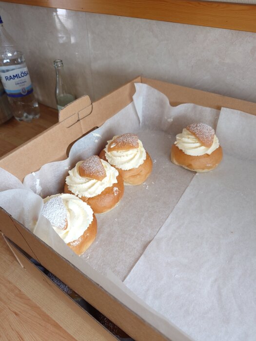 Fyra semlor med grädde och lock pudrade med florsocker i en kartong på ett bord, i bakgrunden syns flaskor.