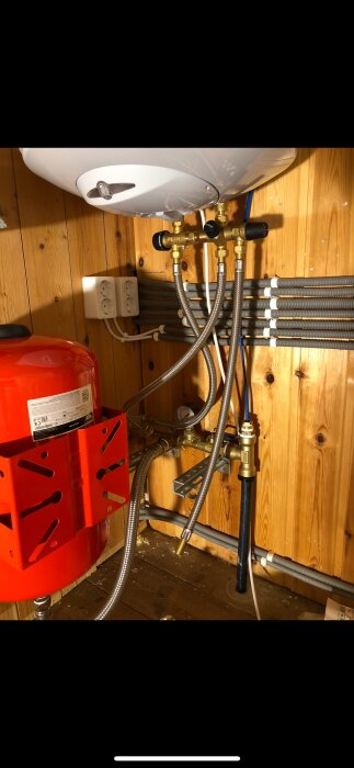 Kompakt vattensystem i fritidshus med röda och vita komponenter, slangar och kopplingar i trävägg. Begränsat utrymme för vattenfilter.