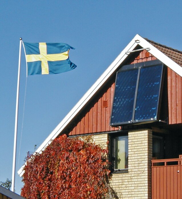 Solfångare monterad på husets tak, nära lodrät med svensk flagga i bakgrunden.