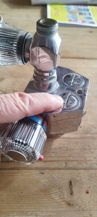 Hand som pekar på gängad plugg på metallventil med vred, liggande på träyta.