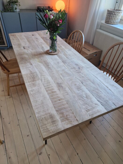 Ljust mangoträbord med rustik yta, dekorerat med en vas med blommor, står i ett rum med trästolar och parkettgolv.
