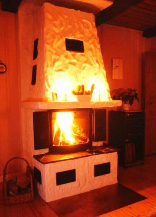 Vit eldstad med brasa och glasluckor i ett vardagsrum, omgiven av ljus och dekor.