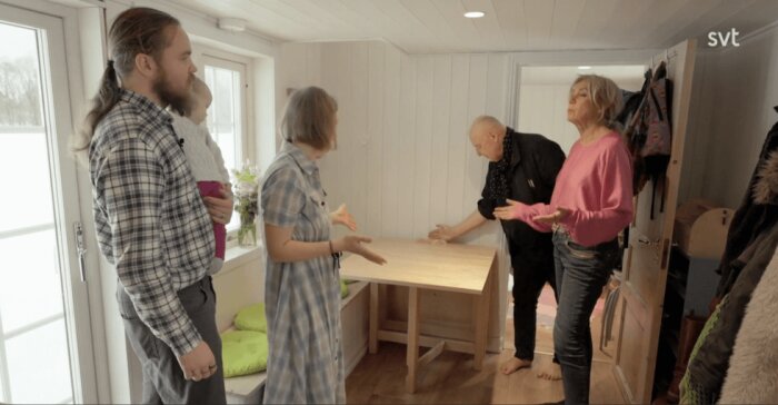 Fyra personer diskuterar runt ett träbord i ett litet, ljust rum med träpanel, en man håller ett barn i famnen.