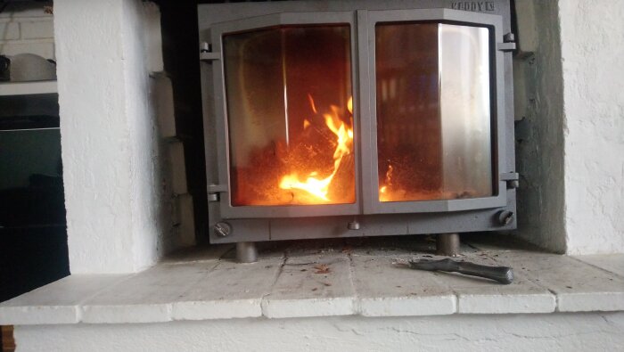 Braskaminens framsida med eld inuti, placerad på oregelbunden yta. En skruvmejsel ligger framför kaminen på en stenyta.