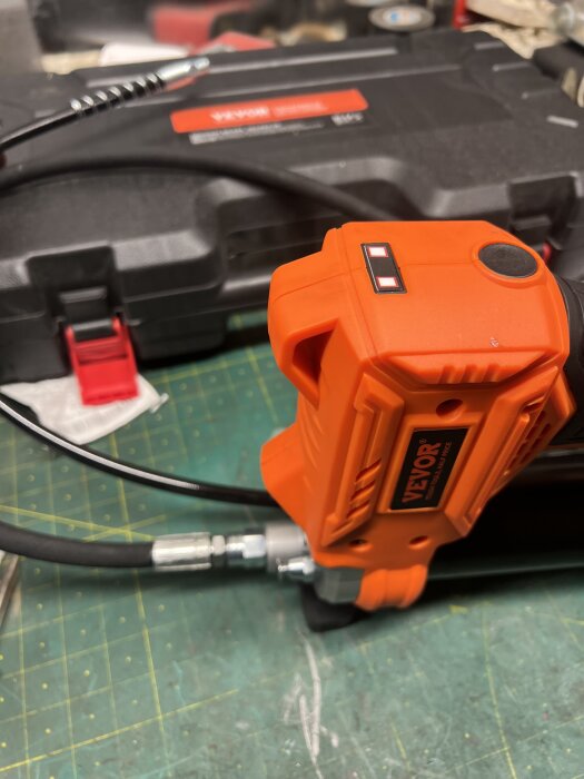 Elektrisk fettspruta av märket Vevor med orange handtag och slang, placerad på arbetsbord med verktygslåda i bakgrunden.