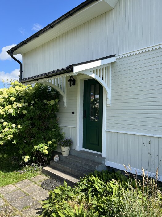 Fasad med vit träpanel och grön dörr, omgiven av trädgård. Taktegel och marksten syns.