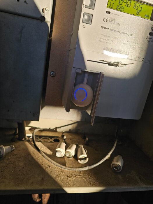 Elmätare från eon med RJ12 uttag, omgiven av flera glödlampor och dammig yta. En HomeWizard P1 är inkopplad.