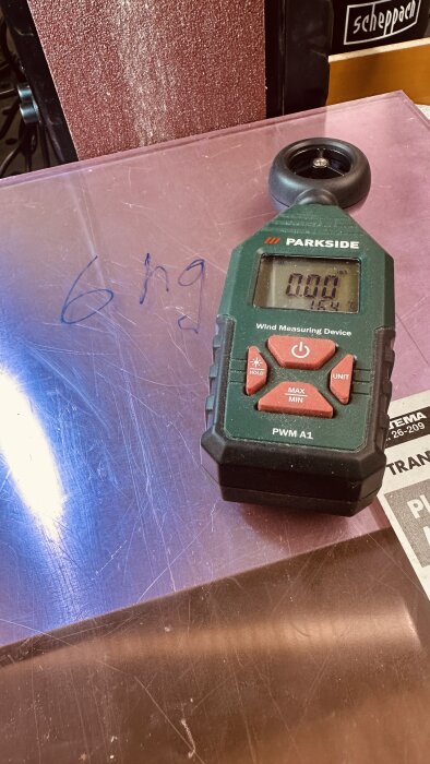 En Parkside vindmätare ligger på en plexiglasskiva med texten "6 hg" skrivet med blått. I bakgrunden syns en Scheppach verktygslåda.