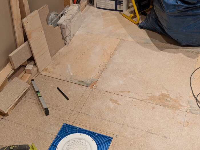 Pågående badrumsrenovering med golvplattor av spån, nivåmätare och verktyg. En blå golvbrunn syns i hörnet av bilden.