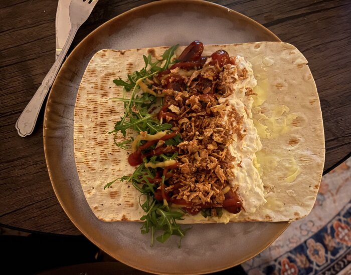 Tunnbrödsrulle med ruccola, kabanoss, räksallad, hemgjort mos, rostad lök, bostongurka, ketchup och senap på tallrik.