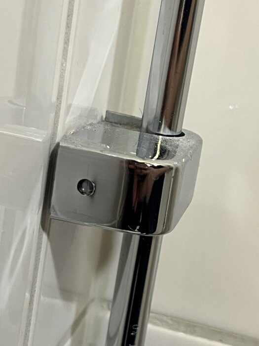 Kromat väggfäste för vattenrör monterat mot en kaklad vägg i badrum, med synligt skruvhål och viss silikon runt kanterna.