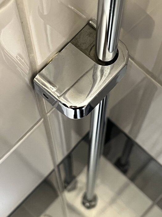 Kromat väggfäste som håller ett vertikalt vattenrör mot en kaklad vägg i ett badrum, del av ett rörsystem.