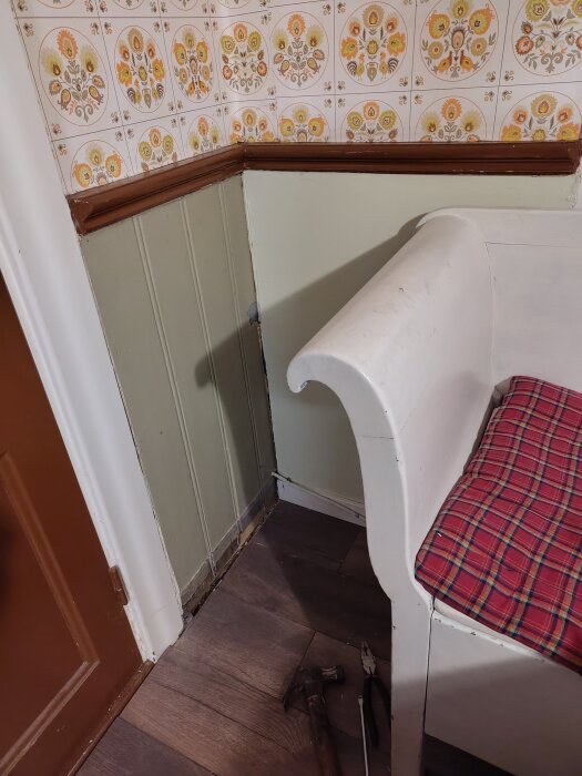 Köksvägg med delvis synlig pärlspontpanel under masonit, blommigt kakel ovanför, vit soffa med röd kudde, verktyg på golvet.