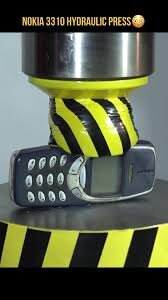 En gammal mobiltelefon, inlagd under ett tryck från en hydraulpress, med svarta och gula varningsränder.