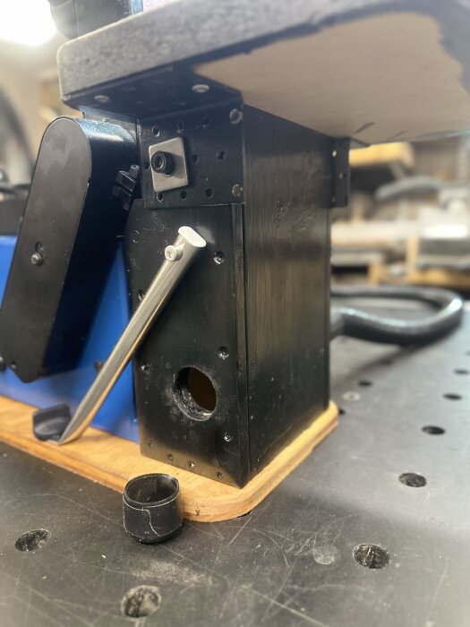 Bilden visar basen av en svart bandslipmaskin med aluminiumstomme och fästen, monterad på en arbetsbänk, med en plugg på golvet.