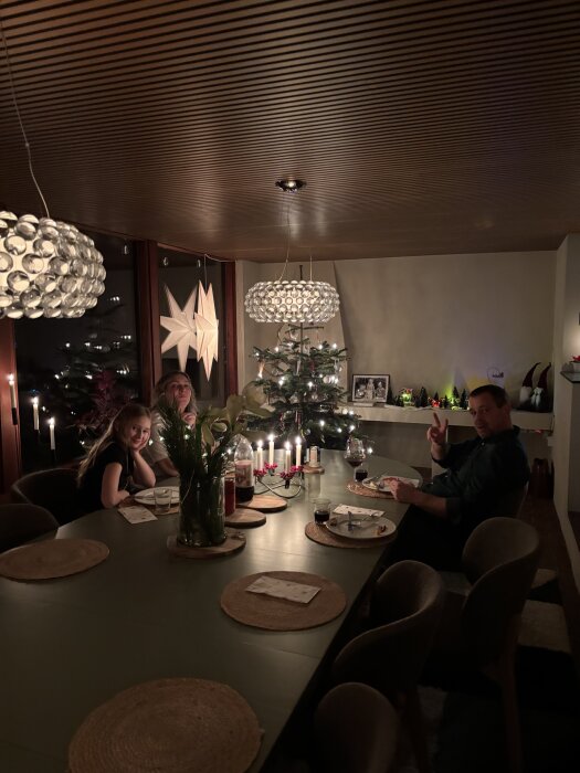 En grupp människor sitter vid ett matbord i ett rum med träpaneler i taket och julpynt, inklusive en julgran och stjärnformade fönsterdekorationer.