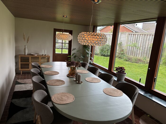 Matplats med lång bord, åtta stolar, taklampa och stora fönster ut mot trädgård. Taket har träpanel.