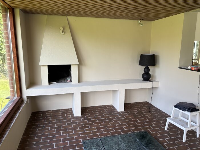 En inomhusvy med en vit öppen spis, en svart lampa på en bänk och ett tegelgolv. En vit trappa står till höger om bänken.