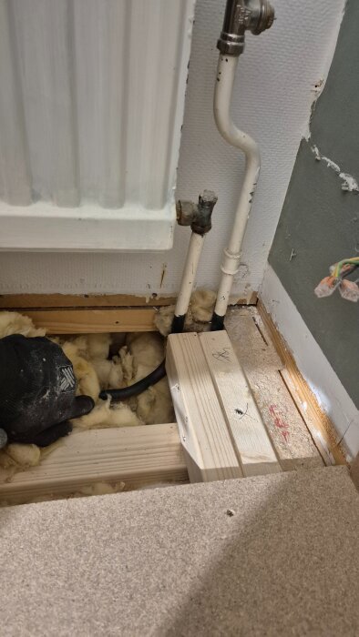 En radiator med kopparrör och isolering synliga genom ett öppet golv, där en hand i arbetshandske rör vid isoleringen.