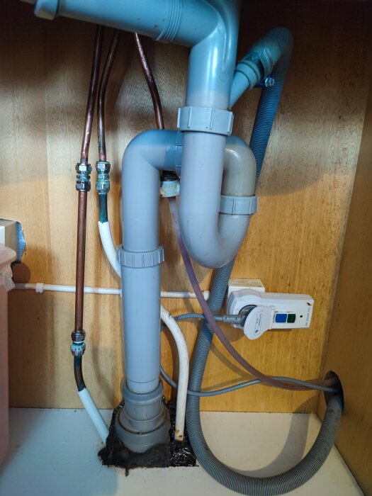 Vattenrör och avloppsinstallationer under diskbänk med olika kopplingar, klämringar och en vattenlås i ett köksskåp.