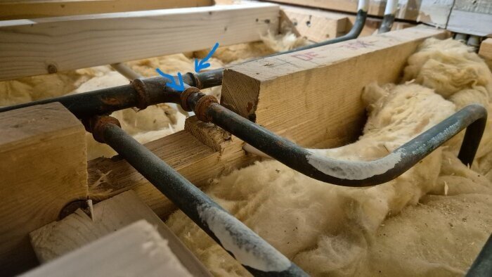 Vattenrör som går genom isolering under ett trägolv, med två blå pilar markerande en skarv eller koppling.