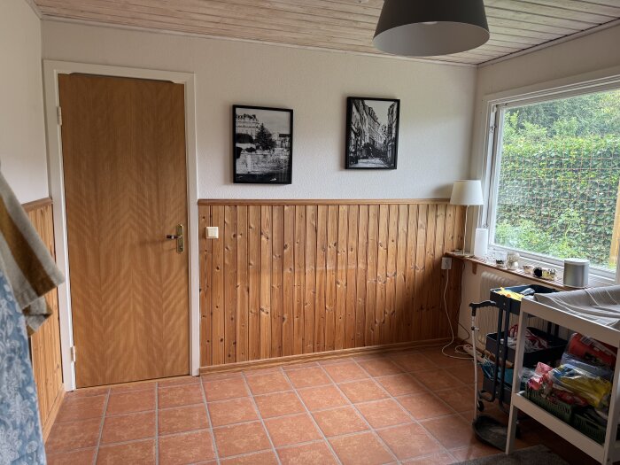 Träpanelvägg, två svartvita tavlor, brun dörr till vänster, fönster till höger med utsikt mot grönska, beige kakelgolv, vit golvlampa.