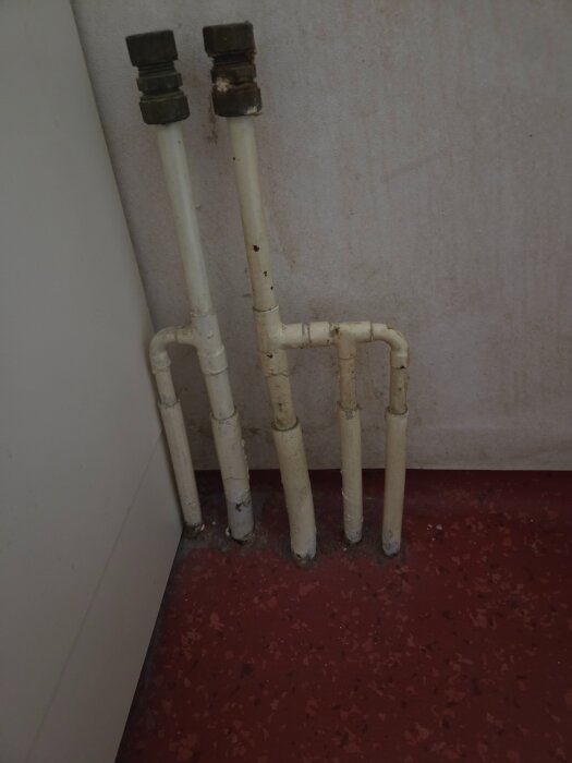 Vita vattenrör som kommer upp från ett rött golv och fortsätter upp längs en ljus vägg, med tre synliga kopplingar längst ner.