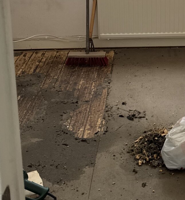 Golv under renovering med delvis borttagen papp och svarta fläckar av lim, borste i bakgrunden. Tecken på möjlig asbestförekomst.