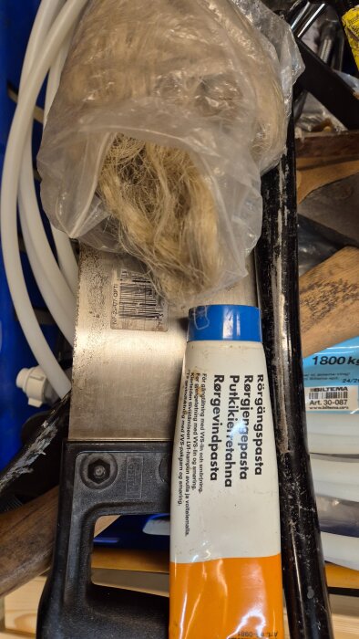 Rördelar och VVS-tillbehör: en tub rörgängspasta, en plastsäck med lin, och en såg, på en blå bakgrund.