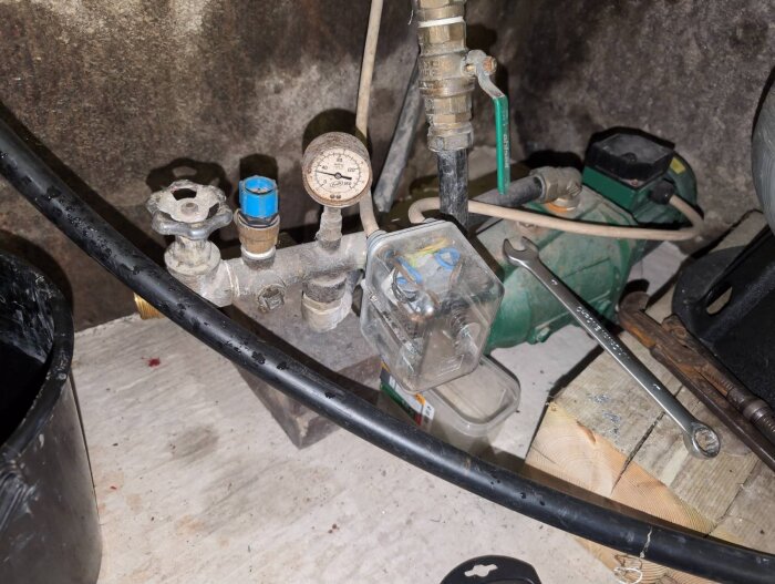 En gammal vattenpumpinstallation i en källare med rör, en manometer och en grön EMS Jet pump 105T, samt verktyg bredvid.