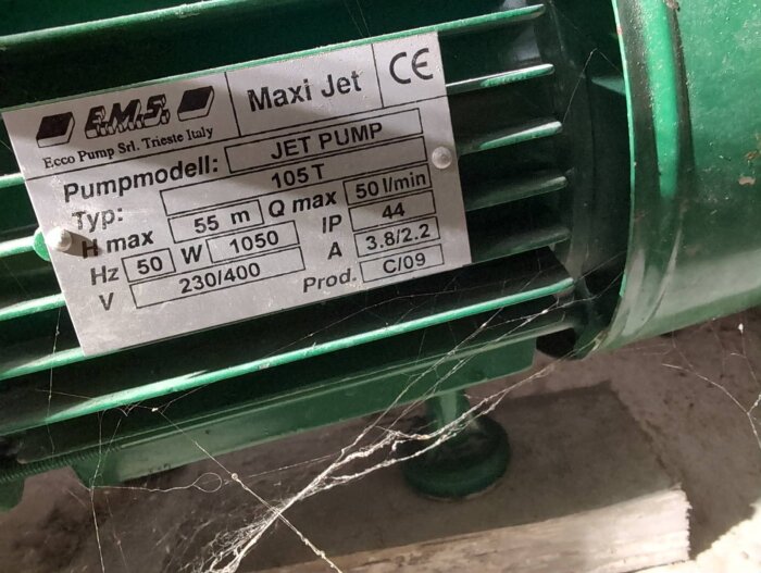 Grön EMS Maxi Jet 105T pump med specifikationer: 55 m, 50 l/min, 1050 W, IP 44, 230/400 V. Synlig spindelväv i bakgrunden.