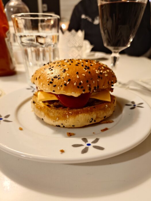 Burger med sesambröd, ost och tomat på en tallrik, omgiven av ett glas vin och ett glas vatten på ett dukat bord.