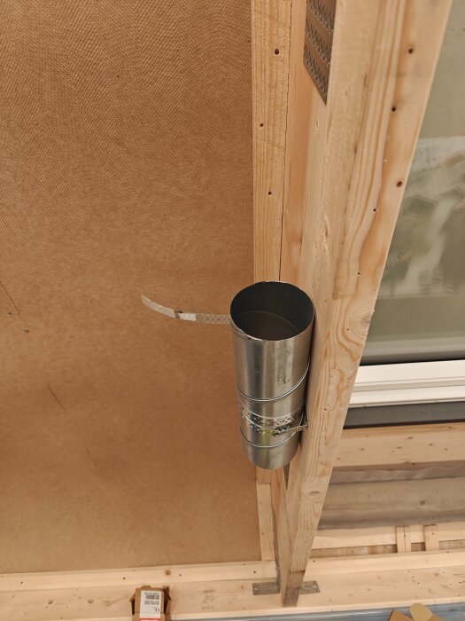 Ventilationsrör i metall fäst vid en träram i taket med hjälp av hålband, sett från sidan.