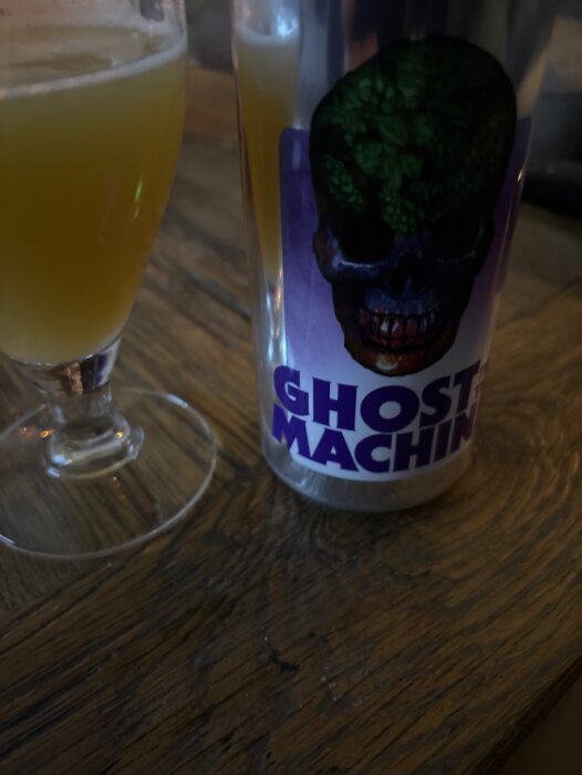 Ett glas med gul dryck och en burk med en färgglad etikett som visar en dödskalle och texten "Ghost Machine" på ett bord av trä.