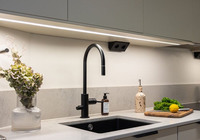 Kök med installerad IKEAS LED-list under överskåp, svart kran, bänkskiva med diskho, och dekorationer som citron och växter.