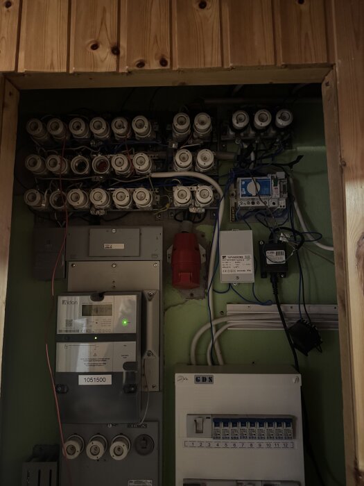 Elcentral med gamla porslinssäkringar, modern elmätare och automatsäkringar monterade på en grön vägg med träpanel ovanför.