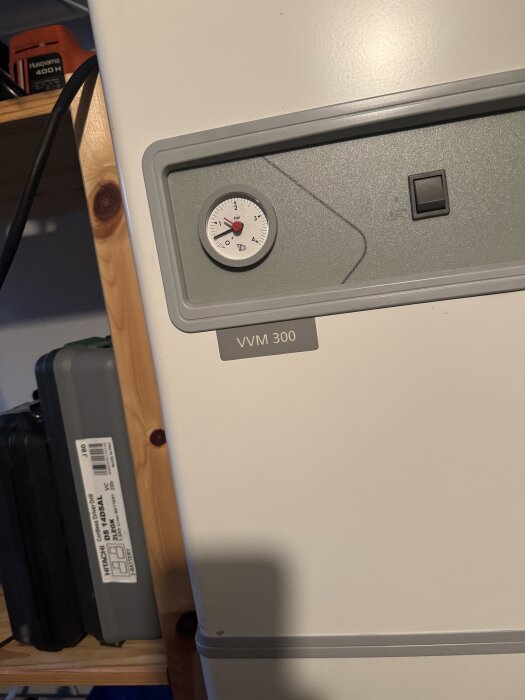 Luftvatten värmepump, modell VVM 300, med manometer, i ett förrådsutrymme. Hylla med verktyg syns i bakgrunden.