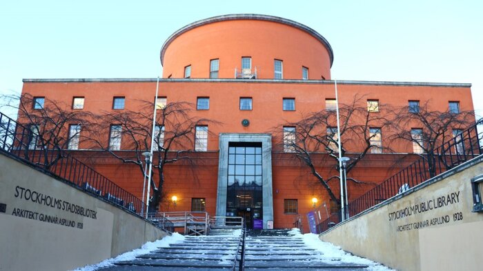 Orange byggnad i Stockholm med trappa och vinterträd; entrén till ett bibliotek med namnet "Stockholms stadsbibliotek" synlig på väggen.